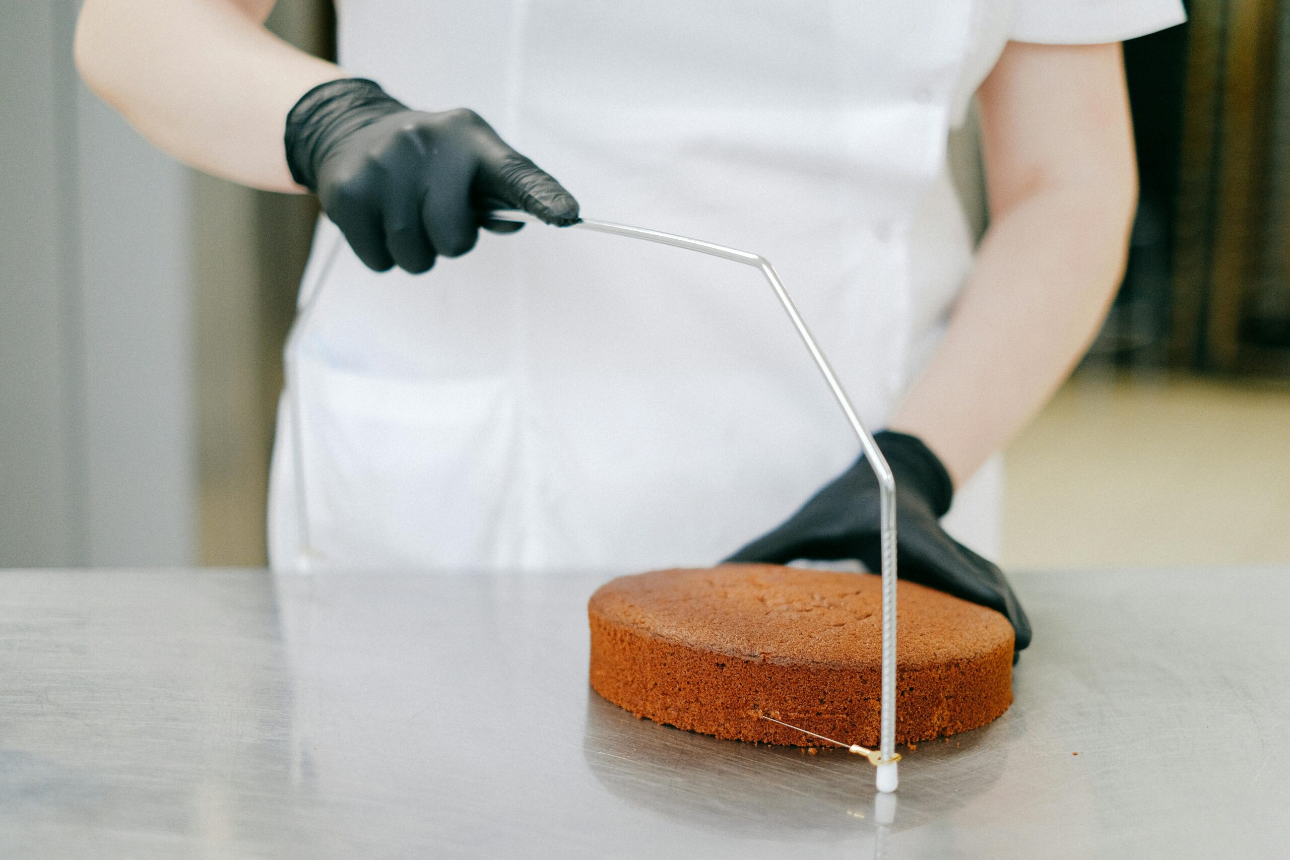 An adult pastry chef slices a round cake with a wire cutter, wearing black gloves in a kitchen.