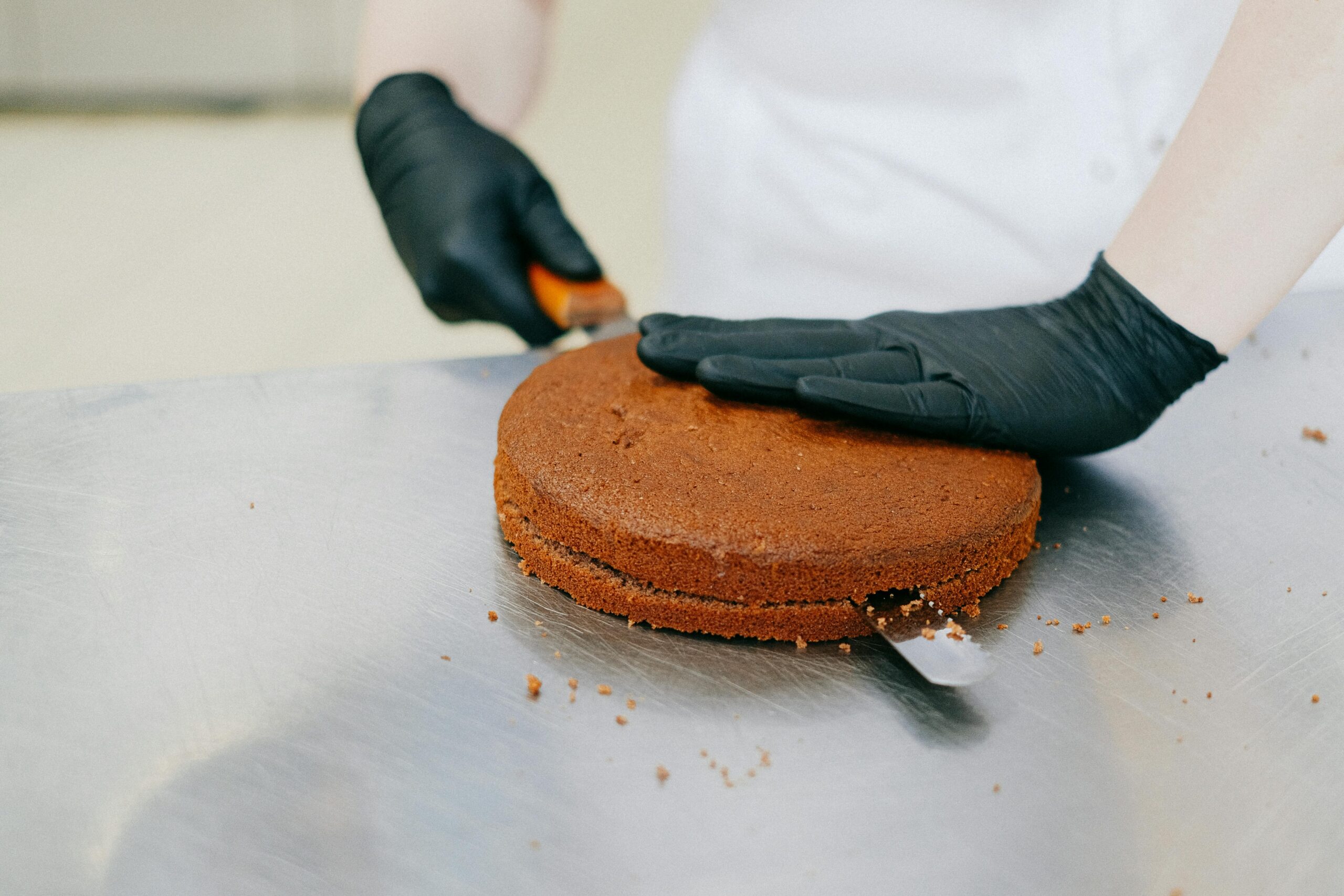 A baker in gloves skillfully slices a chocolate cake with a spatula on a metal table.